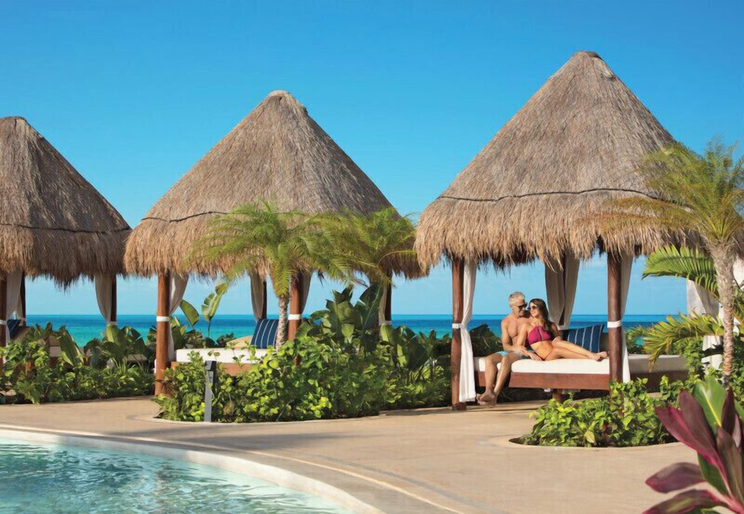 A couple relaxing in a thatched beach cabana beside the pool at Dreams Playa Mujeres, ocean behind