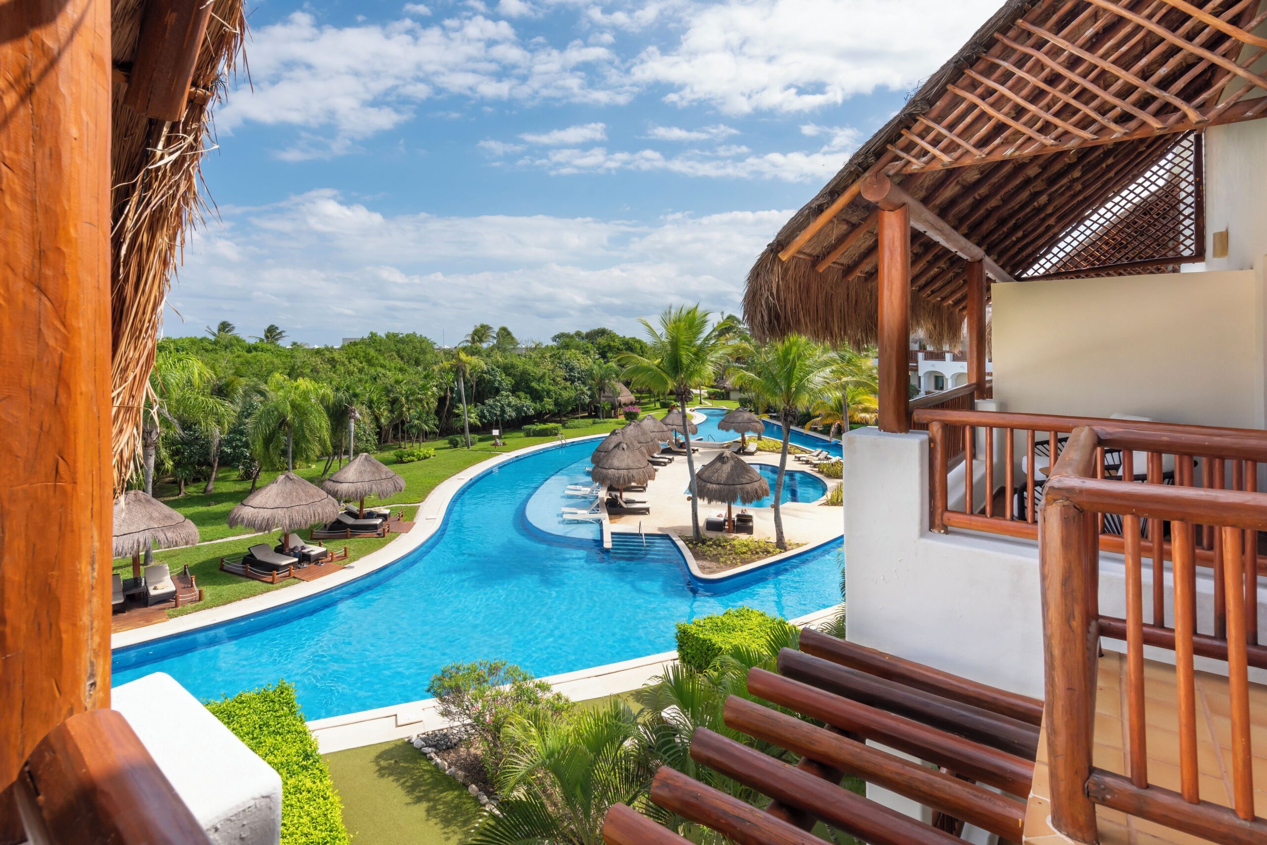 A curving pool lined with palm-thatch palapas seen from a suite balcony at Valentin Imperial Riviera Maya
