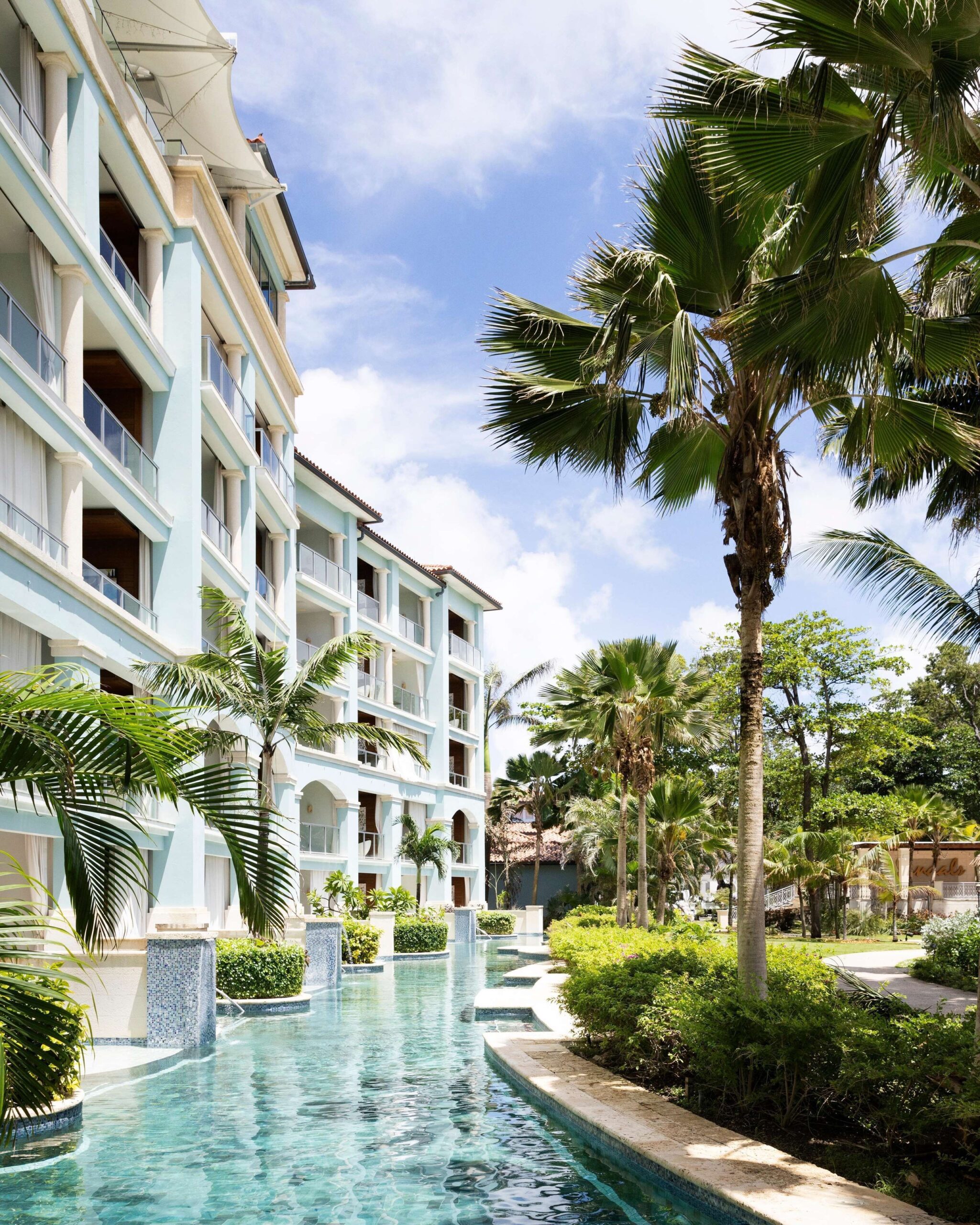 A pale-blue multi-storey suite building with balconies, palm trees, and a pool channel at Sandals Royal Barbados