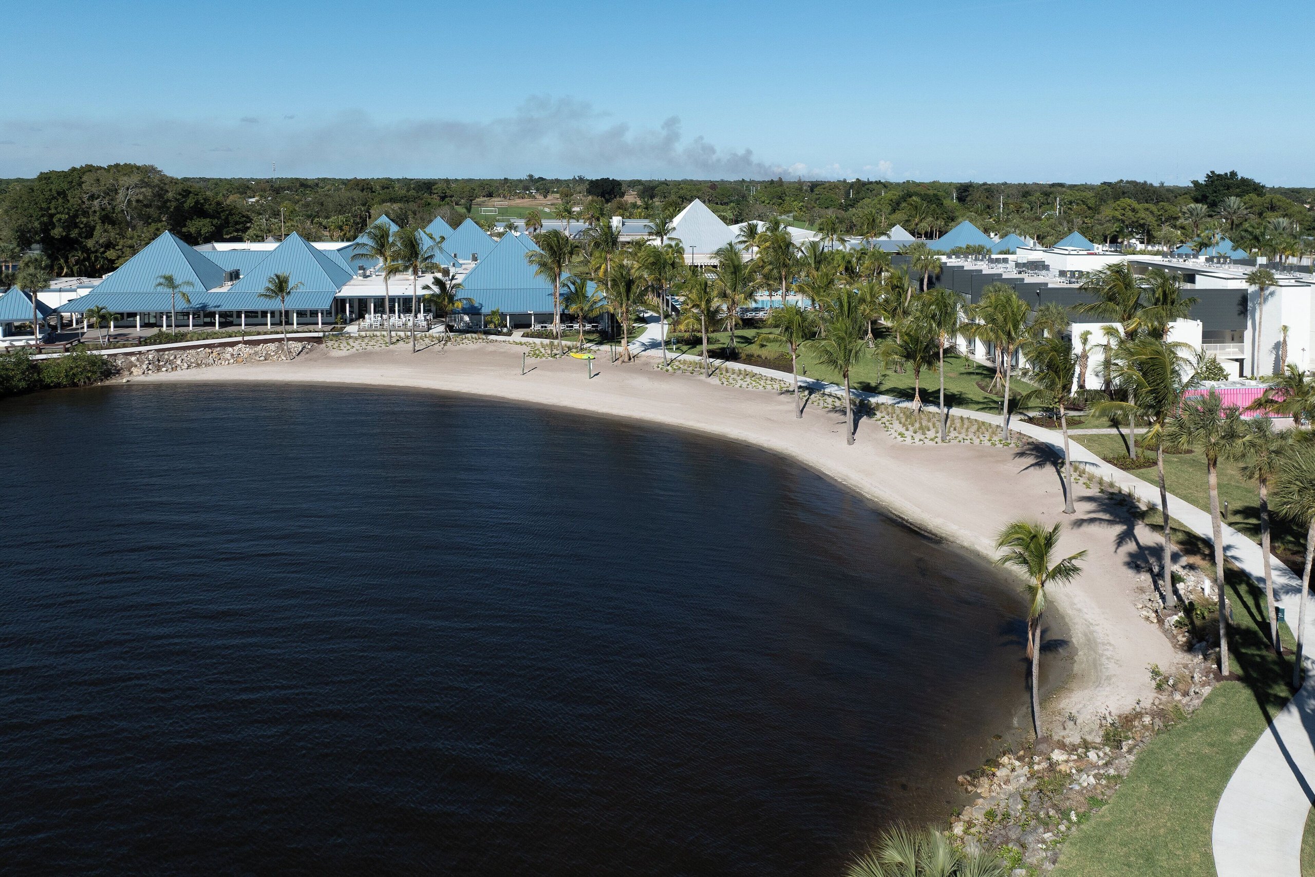 Elevated view of the voco Sandpiper resort's riverside sand beach, palm trees, and blue-roofed buildings on the St. Lucie River