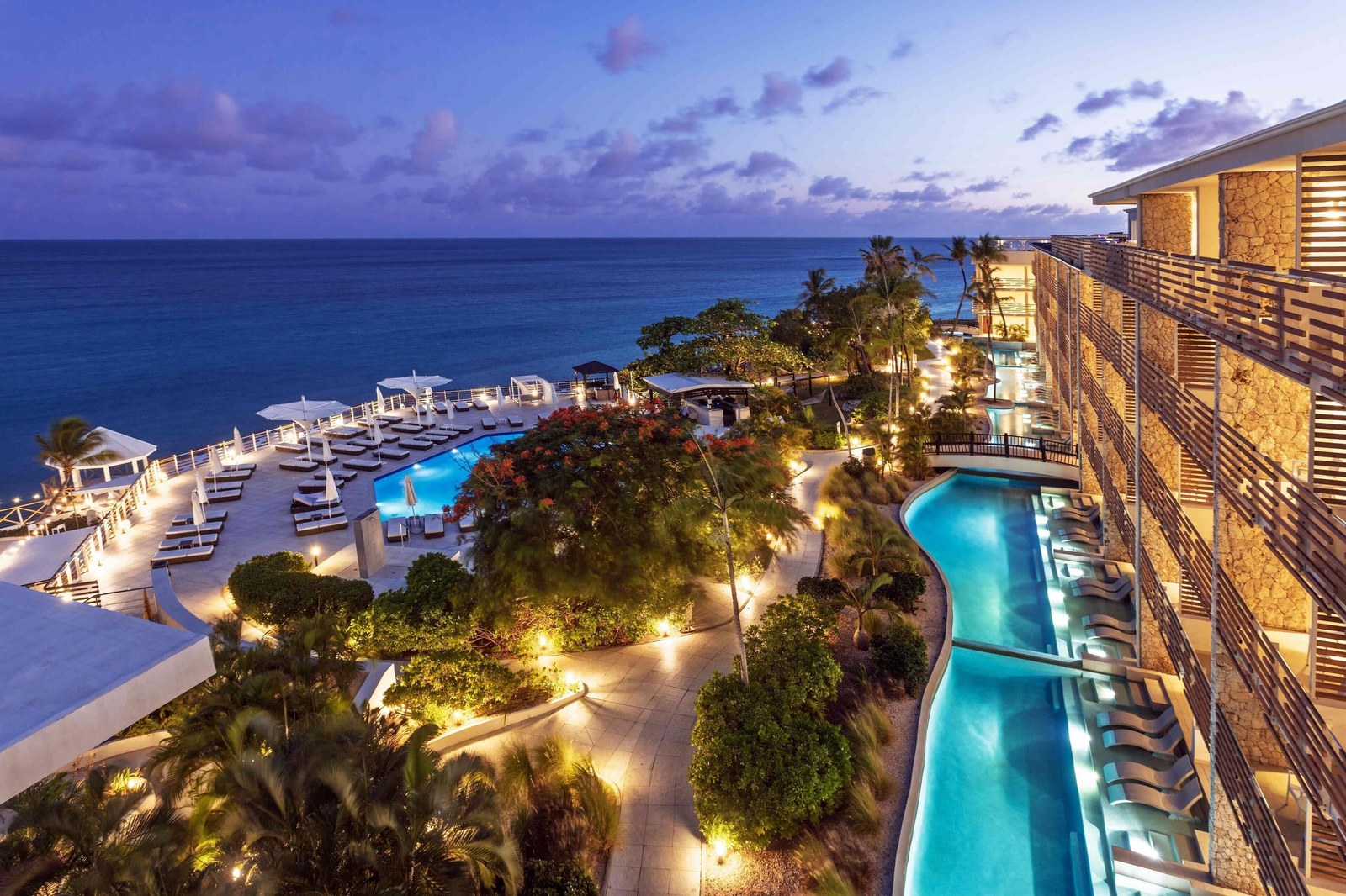 Sonesta Ocean Point suite tower at dusk with the pools, gardens and loungers lit below it